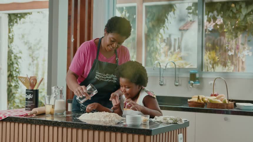 A mother pours water in flour while her daughter smile in a bright kitchen