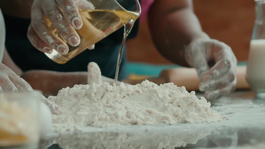 Flour covered hands pour oil into a pile of flour while preparing dough