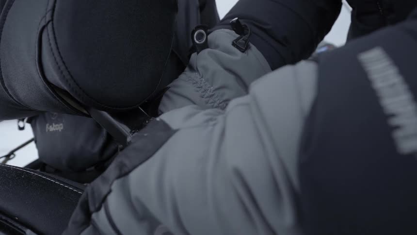 A man wearing heavy winter gear ties a pack to the back of a snowmobile