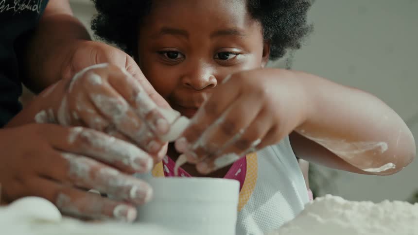 A young child cracks an egg into a bowl with the help of an adult