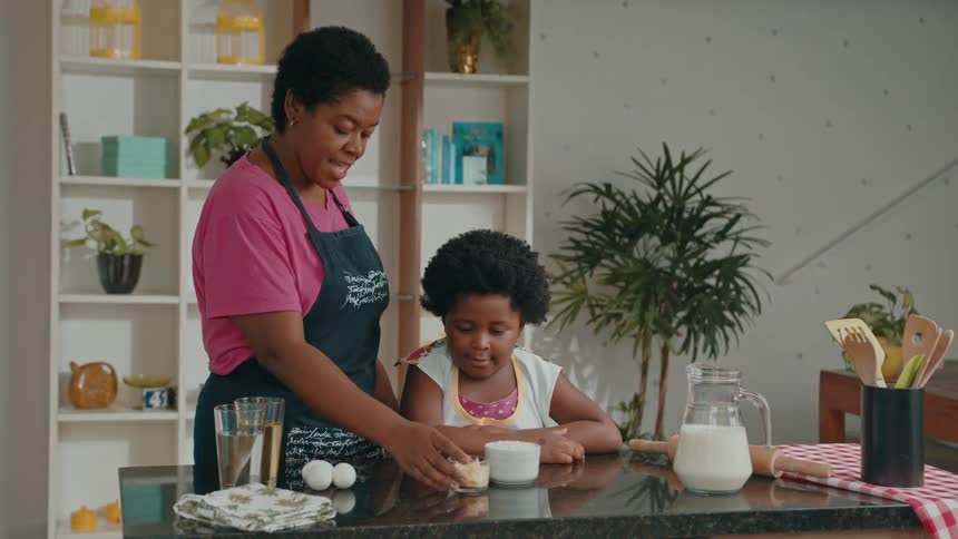 A mother talks to her child while moving some ingredients and eggs in front of the child who is standing at a kitchen table around midday