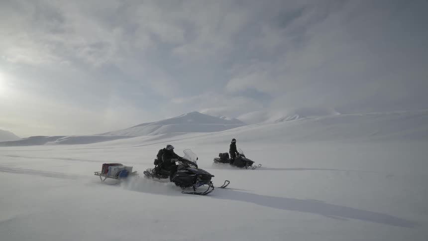 Two people riding snowmobiles ride across the snowy plains