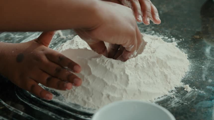 Two pairs of hands are shaping white flour on a dark countertop with a white bowl visible in the foreground