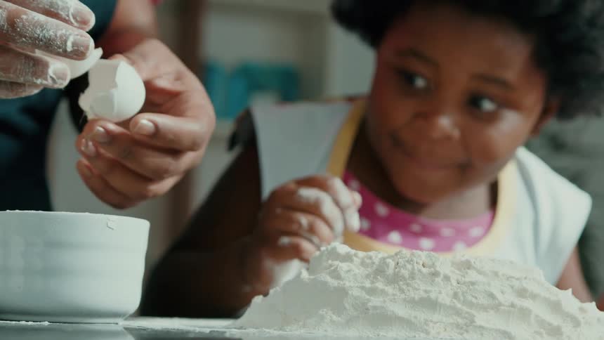 A woman cracks an egg into a white bowl while a child in an apron watches surrounded by piles of flour