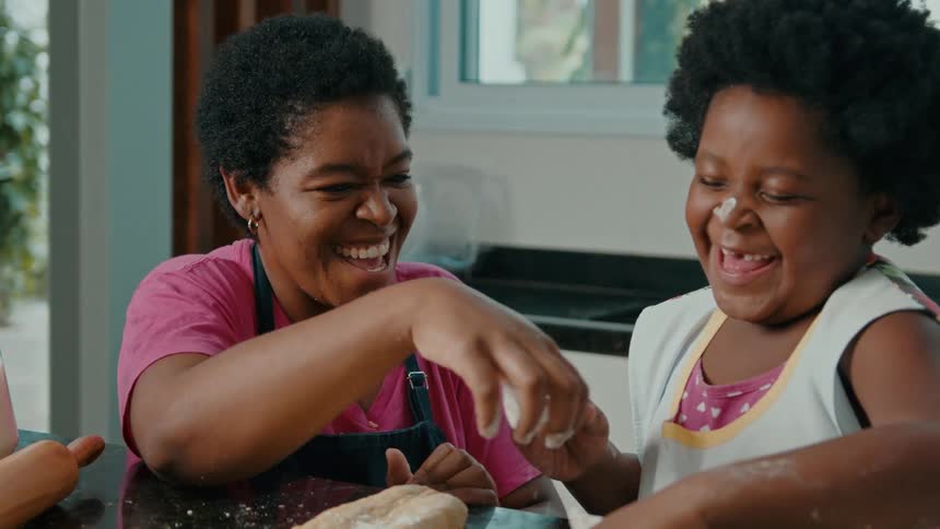 A mother and her daughter with flourdusted arms roll out dough on a kitchen counter preparing bread at home
