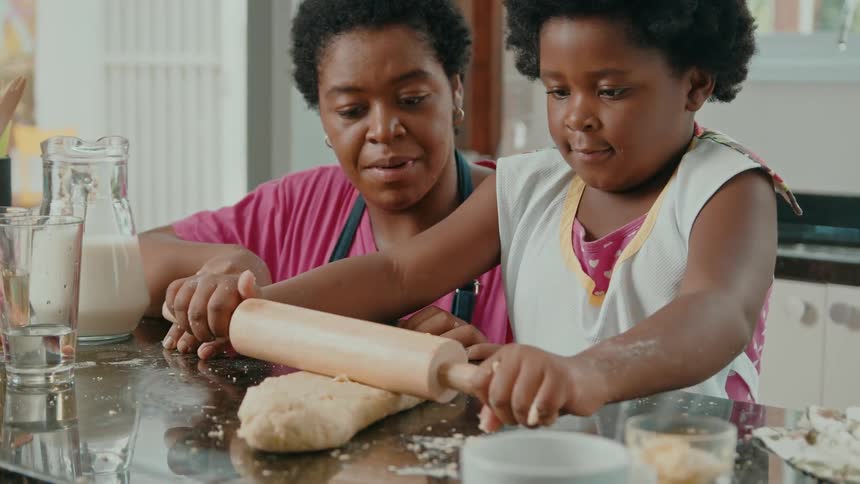 A woman watches her child with excitement as she rolls dough on the countertop with a rolling pin around midday