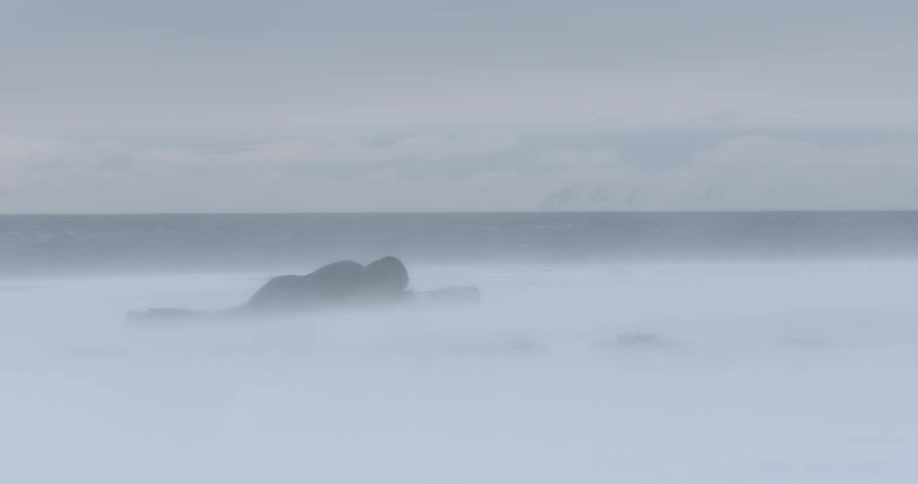 A photographer lies on the ground in a frozen landscape on a windy day