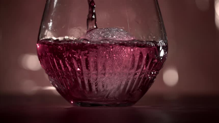 Close up of a glass cup with ice placed on a surface being filled with a red drink