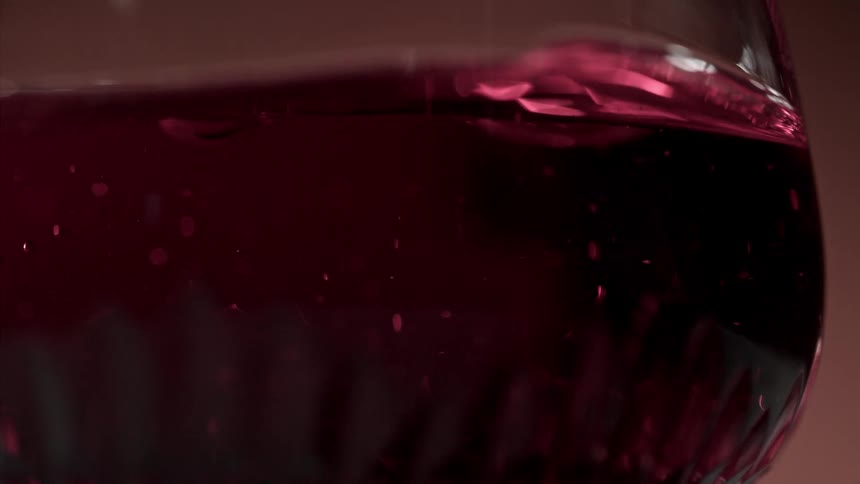 A dark red liquid with tiny bubbles swirls inside a faceted glass captured in closeup against a dimly lit backdrop