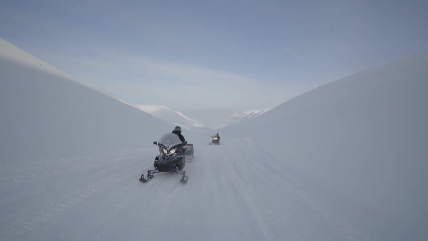 A couple of snowmobilers pulling wagons behind them driving through a valley of snow