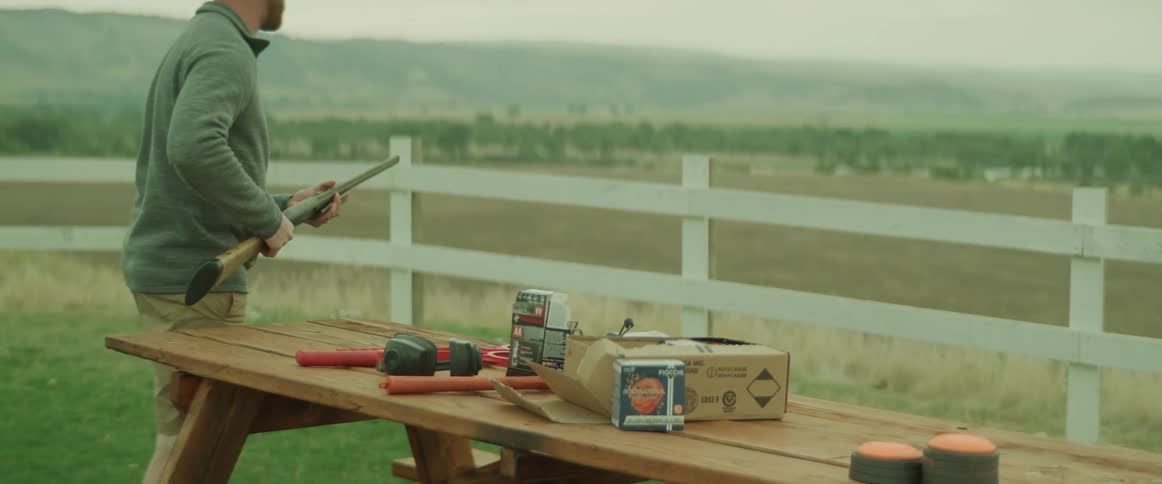 Man picks up a rifle from a picnic table that also contains ammunition and walks toward a fence