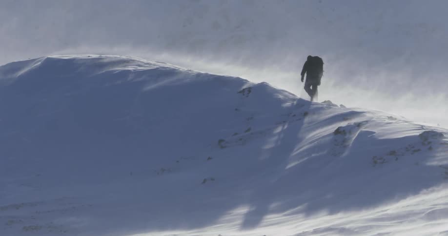 A view of a snow hill and the wind blowing it and a man walking on the peak