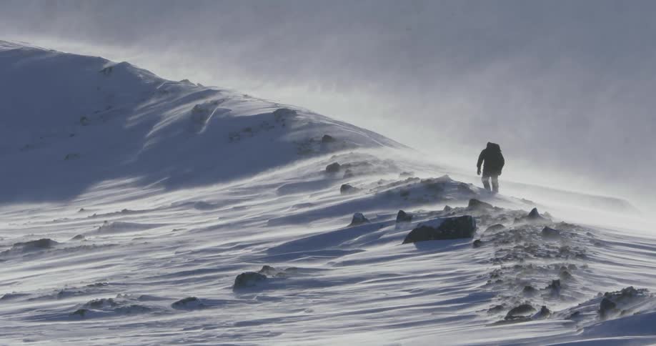 A man is hiking along a snow dune on a windy day