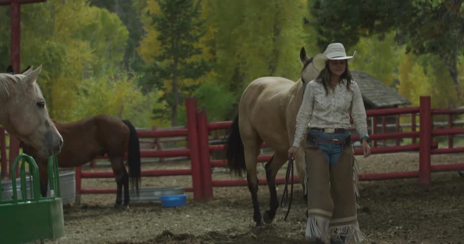 A cowgirl in chaps walks through a dusty corral leading a light colored horse past other horses and a feeding trough with autumn trees and a rustic building in the background