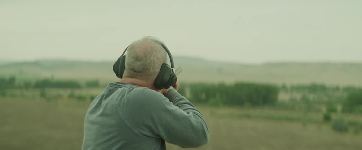 A man wearing earmuffs aims a shotgun across an open rural landscape