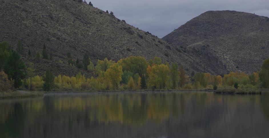 A calm river reflects autumn trees and hills under a cloudy sky