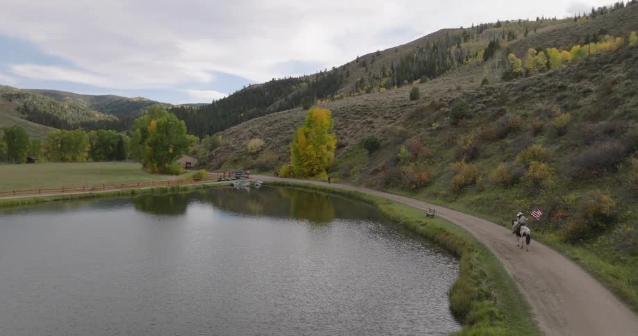 A person on horseback rides along a dirt road beside a calm lake in a hilly fall landscape