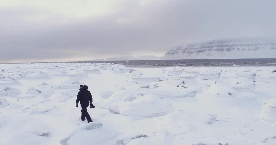 A man with a snow suit carrying a camera is walking over bumpy snow with water in the background