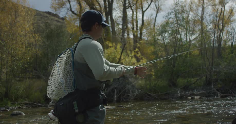 An angler stands mid river in fly fishing gear vest cap sunglasses and net casting into gently rippling water surrounded by autumntinted trees