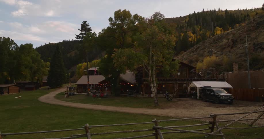 A rustic lodge sits in a wooded valley with autumn foliage and glowing string lights