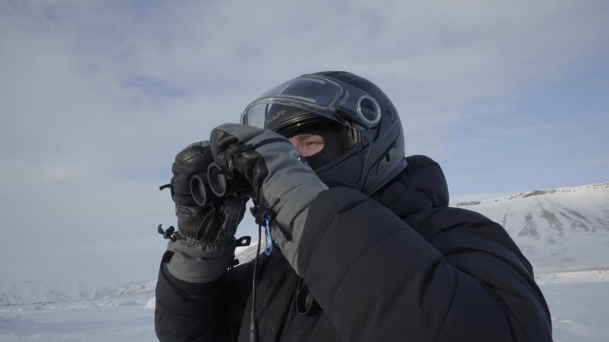 A man wearing a helmet and winter gear looks through binoculars