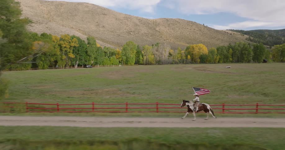 Woman carrying the us flag while riding a horse along a riverbank with mountains in the background