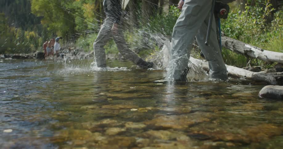 Three men walk over a log from the forest into a stream heading upstream while holding fishing gear and carrying backpacks at midday