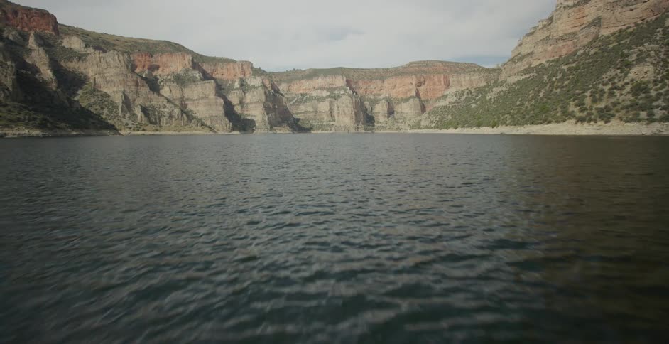 A low aerial shot flies quickly across the surface of a wide river moving toward the base of a massive rocky canyon