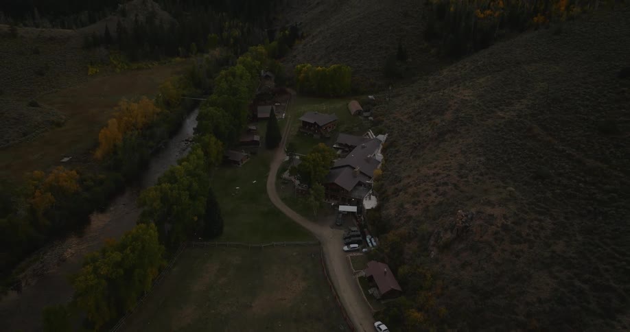 An aerial view shows a collection of buildings nestled in a valley surrounded by a river and hills with a path leading through the property