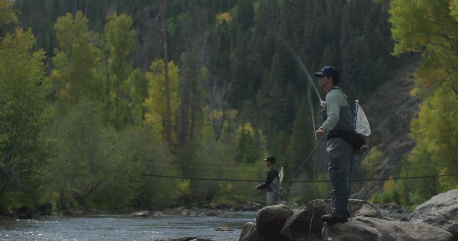 Two people are fly fishing in a scenic river surrounded by trees and mountains