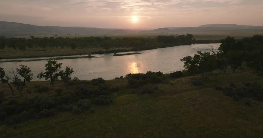 A calm river winds through grassy plains as the sun sets behind distant hills