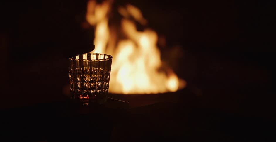 A glass of drink and cigar are silhouetted in front of a glowing fire at night