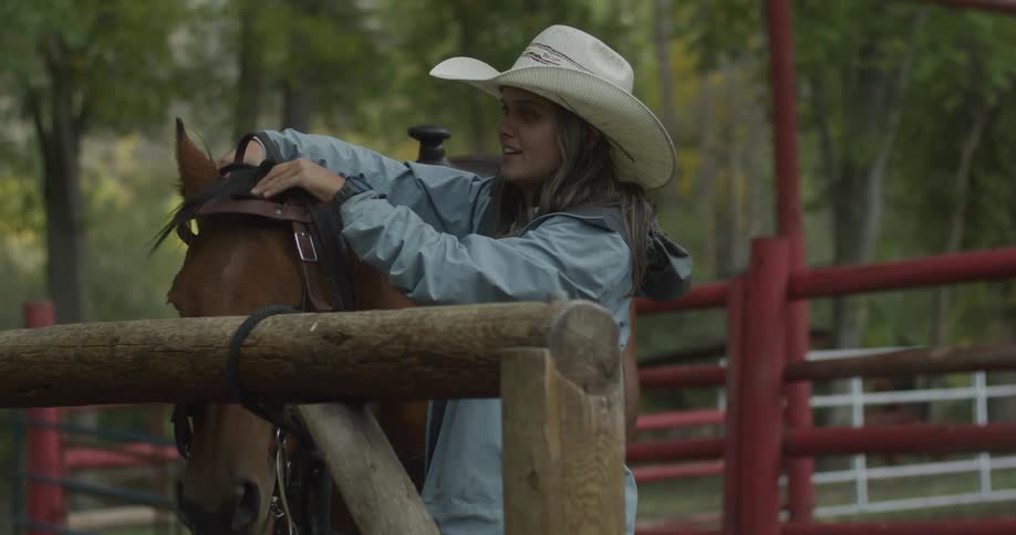 Woman in hat with happy expression putting leather ties on a brown horse