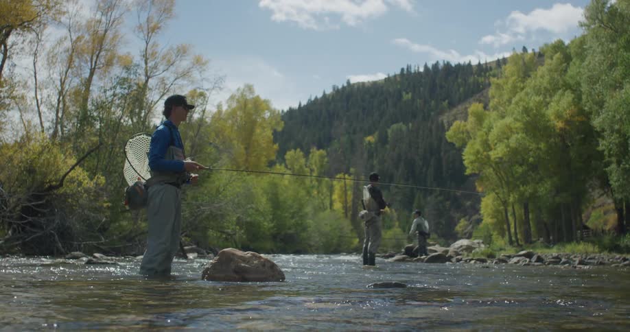 People fly fishing in a shallow forest river surrounded by colorful autumn trees and hills
