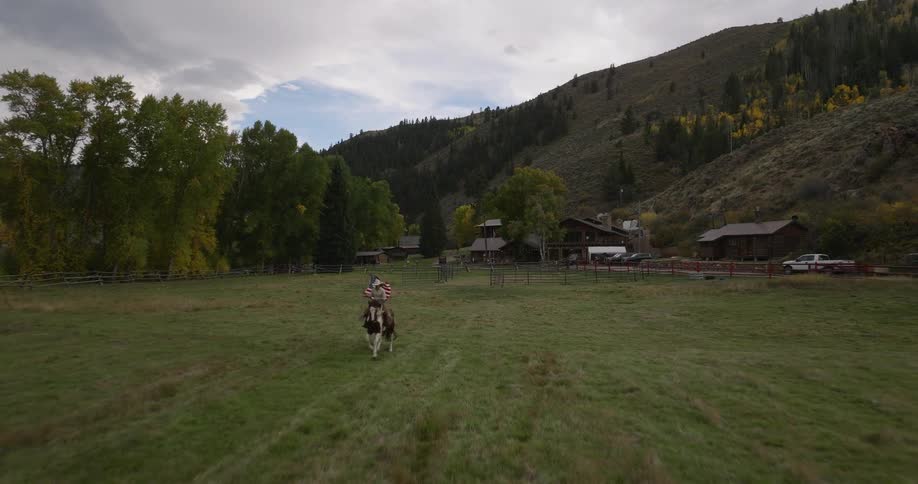 A person on horseback carrying an american flag rides across a grassy field towards a rustic ranch nestled in a mountain valley during autumn