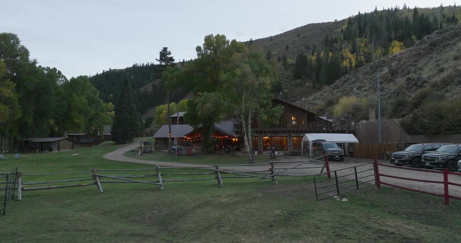 An aerial shot captures a rustic lodge nestled in a valley during autumn with several vehicles parked on a dirt road and guests gathered outdoors under strings of lights