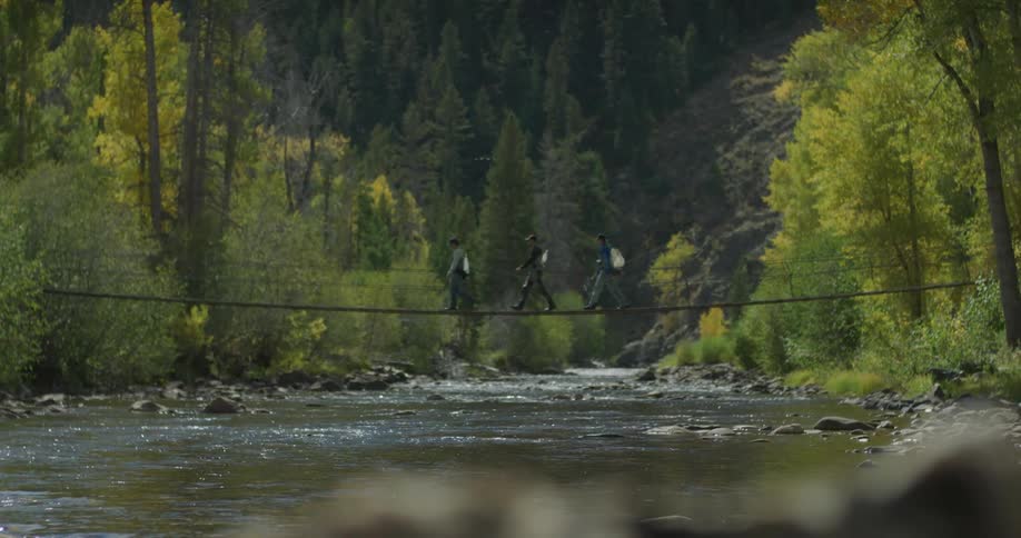 Three hikers traverse a narrow suspension bridge over a forest stream