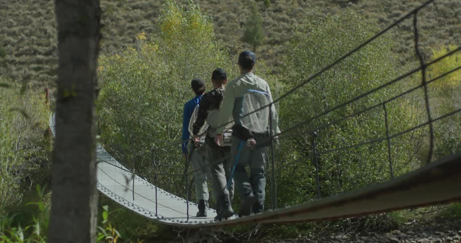 A group of hikers walks across a narrow suspension bridge over a forest stream