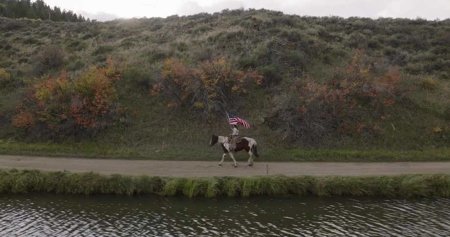 Person carrying the united states flag while riding a horse in a natural setting by a lake