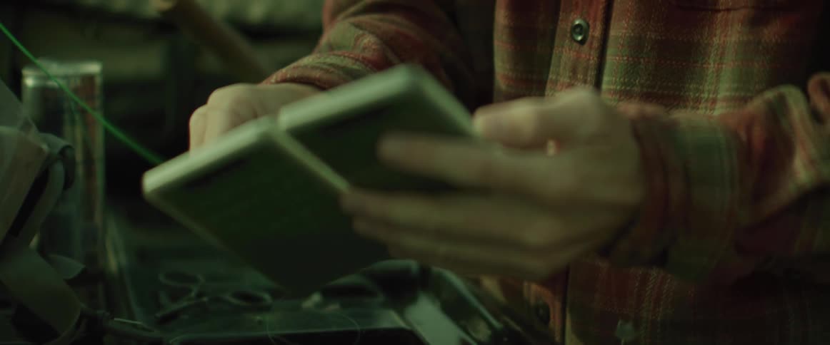 A person in a red and black plaid shirt opens a small case of fishing hooks on a bench
