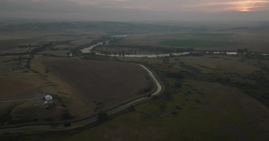 A winding river cuts through patchy farmland and distant hills under a cloudy sunset sky