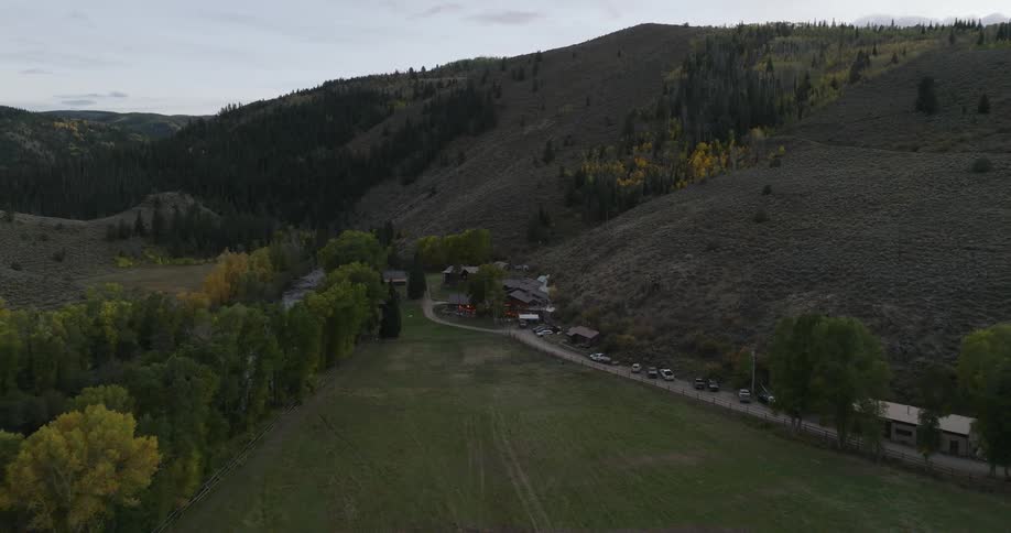 An aerial view shows a sprawling ranch nestled in a valley with colorful autumn trees surrounded by forested mountains under a muted overcast sky