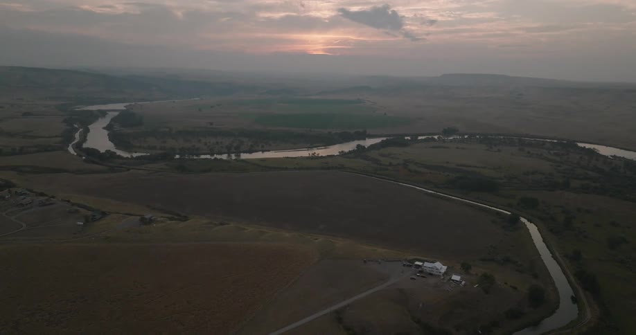 A rural landscape with a winding river and distant hills is seen at sunset from an aerial view