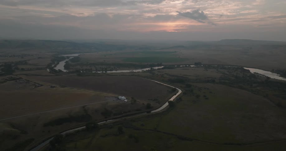 Meandering river winds through golden fields and rolling hills under a glowing sunrise sky with mist softening the distant horizon