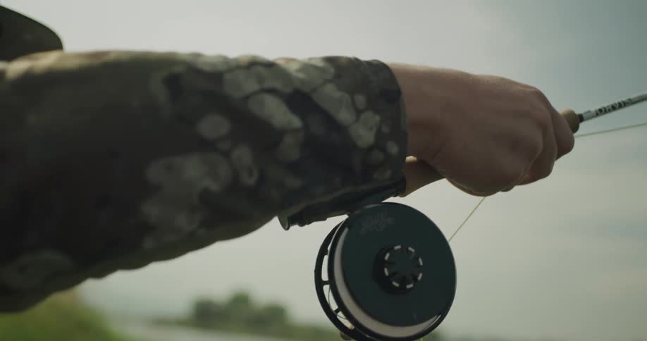 A person fly fishing holding a fishing rod and reel near a riverbank