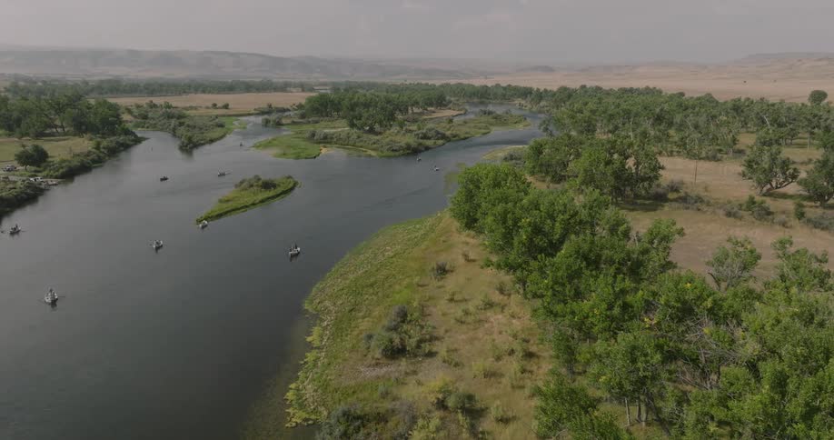 Several small boats navigate a winding river surrounded by lush trees and dry plains under a clear sky