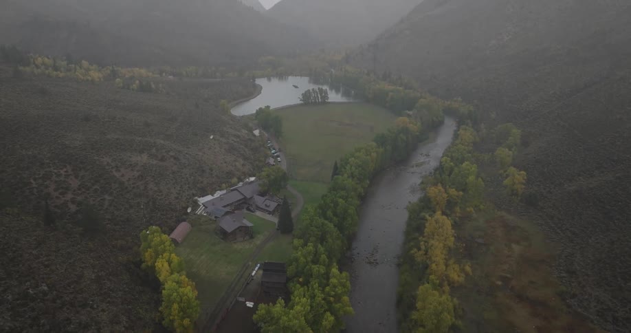 An aerial view shows a winding river alongside a large pond surrounded by hills and trees with autumn foliage and a cluster of buildings with dark roofs nestled by the water all under a cloudy sky