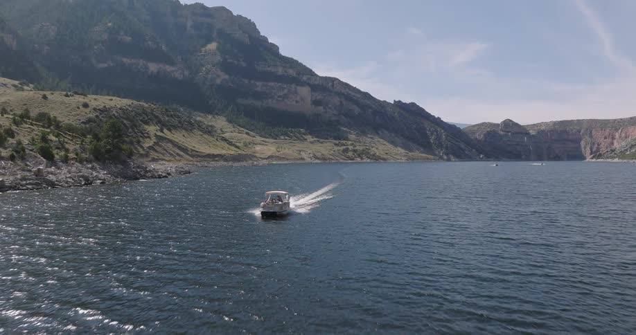A motorboat with people aboard creates a wake as it travels across a large body of water surrounded by imposing rocky mountains under a clear sky