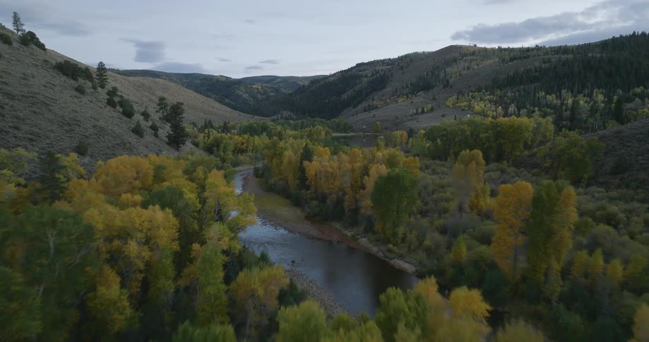 An aerial view follows a winding river through a vast mountain valley dotted with colorful autumn trees under an overcast sky