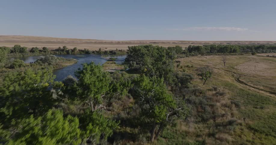 A small wooden cabin sits near a calm river in an open grassland under clear skies